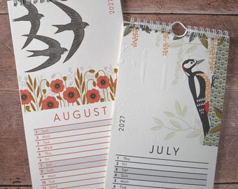 May include: Two 2027 wall calendars with spiral binding. The August page features three black swallows flying above red poppies. The July page displays a woodpecker perched on a tree branch with green leaves and a textured white background.