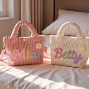 May include: Two corduroy tote bags, one pink and one off-white, with matching handles and shoulder straps. The pink bag has the name "Mia" and the off-white bag has the name "Betty" embroidered on them in colorful letters, with flower accents.