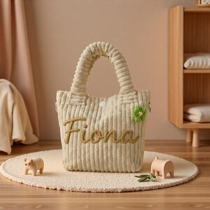 May include: A beige corduroy tote bag with two top handles and a white snap closure. The front features the name Fiona embroidered in a cursive yellow font and a small green crocheted flower accent attached to the upper right side of the bag.