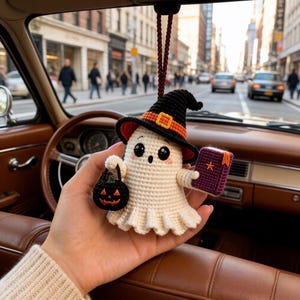 May include: A crocheted ghost ornament featuring a black witch hat with an orange band, holding a small black jack-o'-lantern bucket in one hand and a purple book with an orange star emblem in the other, suspended by a brown braided cord.