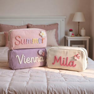 Pode incluir: Três nécessaires de veludo cotelê em rosa, lavanda e creme. Cada bolsa apresenta um nome em uma cor contrastante, com detalhes florais ou decorativos. A bolsa superior diz "Summer", a do meio "Vienna" e a inferior "Mila".