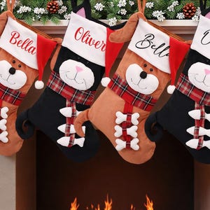 Op de afbeelding: Vier kerstsokken in de vorm van dieren, twee lichtbruine hondensokken en twee zwarte kattenkousen. Elke sok heeft een witte boord met een naam, een rode kerstmuts, een geruite sjaal en witte bot- of visvormige accenten aan de voorzijde.