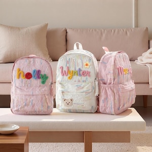 May include: Three backpacks with a textured, multi-colored pastel pattern. The left bag features the name "holly" in colorful letters. The center bag displays the name "Wynter" with a small flower and a bear patch. The right bag shows the name "Maeve".