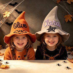 May include: Two crocheted witch hats, one orange with the name Olive and a black bat, and one gray with the name Clara and a black spiderweb. The hats feature a pointed crown and a wide, wavy brim, crafted with a textured knit pattern.