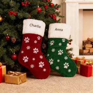 May include: Two Christmas stockings, one deep red and one dark green, each featuring a white faux fur cuff embroidered with the names Charlie and Anna respectively. The stockings are decorated with white embroidered paw prints and snowflakes.