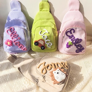 May include: Four corduroy sling bags in pastel shades: light purple, green, and pink. Each bag features an embroidered name on the front, with a flower, car, or grape design. The bottom bag is beige with a hedgehog design.