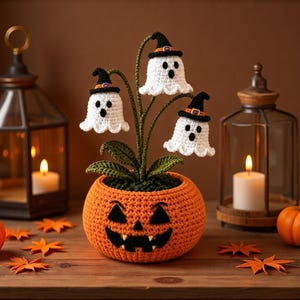 Könnte beinhalten: Eine Häkelpflanze mit drei weißen Geisterfiguren, die schwarze Hexenhüte tragen und aus einem runden, orangefarbenen, kürbisförmigen Topf mit einem schwarzen Jack-o'-Lantern-Gesicht wachsen. Die Pflanze hat grüne Stängel und Blätter.