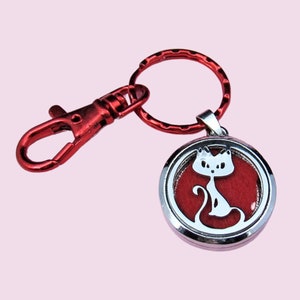 May include: A keychain featuring a red metal lobster clasp and a circular ring attached to a silver-toned circular pendant. The pendant contains a cutout silhouette of a cat against a solid red background, creating a distinct contrast.