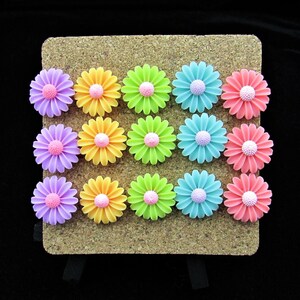 May include: A collection of fifteen flat-backed flower embellishments arranged in a grid of three rows and five columns. The flowers feature vibrant purple, yellow, lime green, light blue, and pink petals with textured, light pink centers.