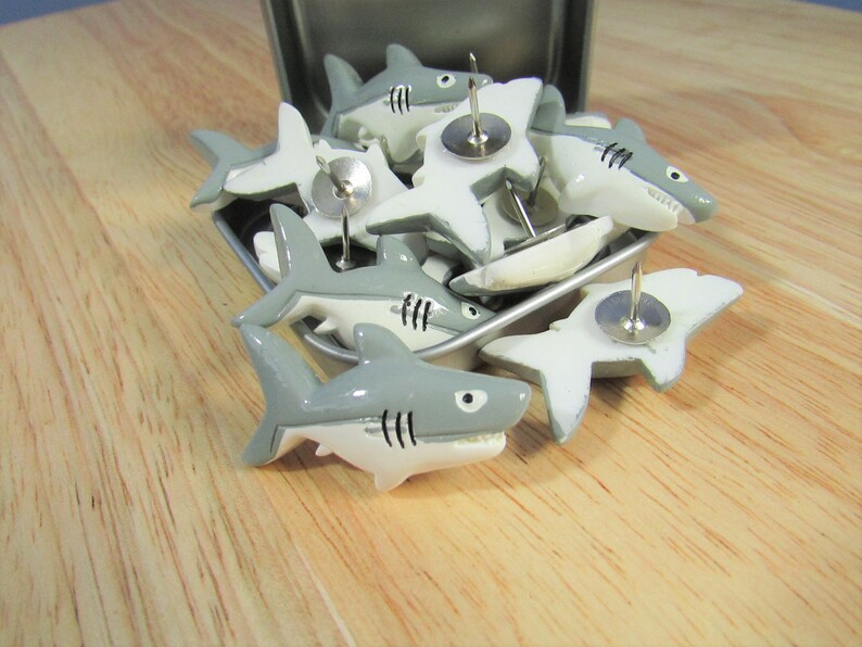 15 Great White Shark Push Pins. They are about an inch long and 1/2 inch high. They come in a metal box with window.