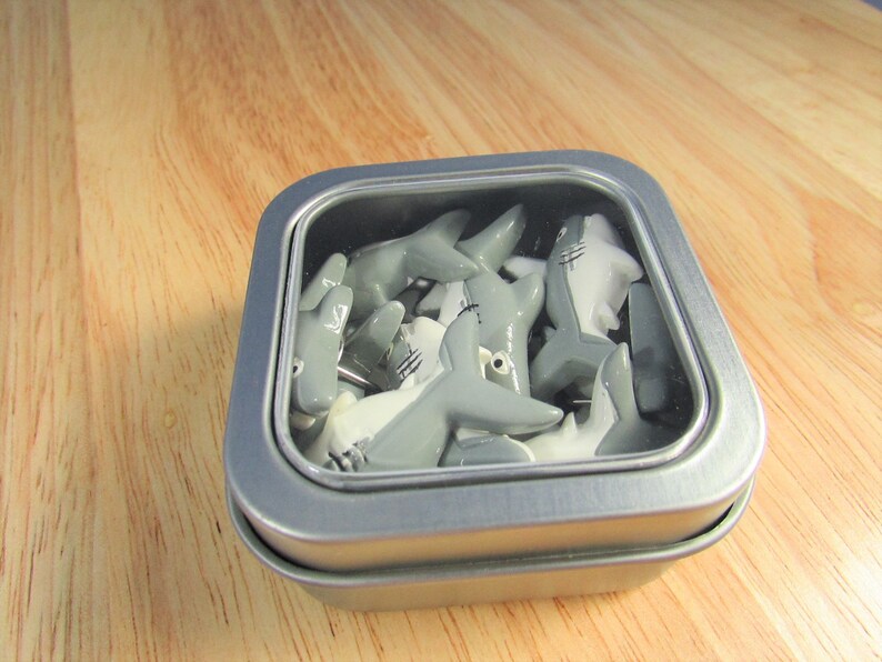 15 Great White Shark Push Pins. They are about an inch long and 1/2 inch high. They come in a metal box with window.