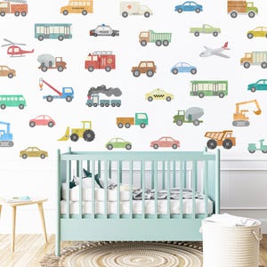 May include: A nursery wall decorated with colourful vehicle decals, including a school bus, police car, fire truck, ambulance, taxi, and construction vehicles. The decals are arranged in a random pattern on a white wall. A cot is in the foreground.