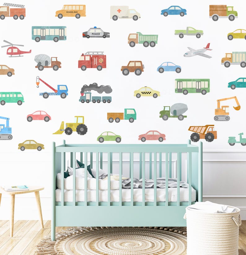 May include: A nursery wall decorated with colourful vehicle decals, including a school bus, police car, fire truck, ambulance, taxi, and construction vehicles. The decals are arranged in a random pattern on a white wall. A cot is in the foreground.