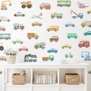 May include: A white wall decorated with colourful vehicle decals, including a school bus, a police car, a fire engine, a helicopter, a plane, a train, and a scooter. The decals are arranged in a random pattern. There is a white shelf with a book titled "Home Philosophy" on top of it.