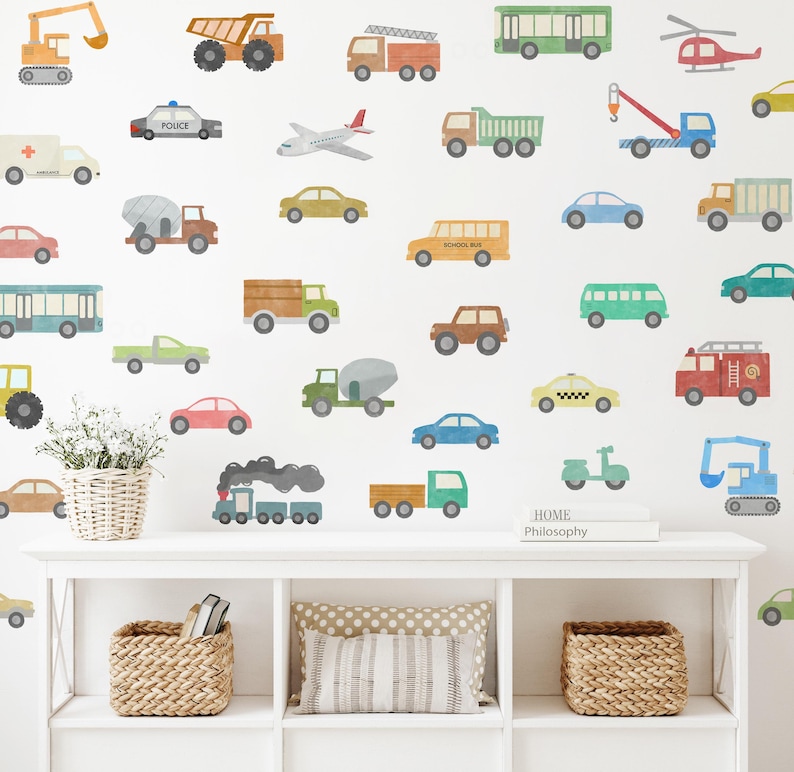 May include: A white wall decorated with colourful vehicle decals, including a school bus, a police car, a fire engine, a helicopter, a plane, a train, and a scooter. The decals are arranged in a random pattern. There is a white shelf with a book titled "Home Philosophy" on top of it.