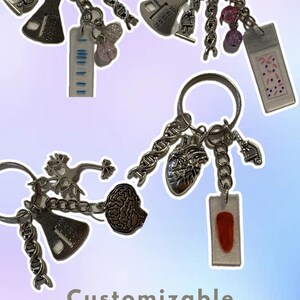 Op de afbeelding: Zilverkleurige, op biologie geïnspireerde sleutelhangers met diverse bedels, waaronder een DNA-streng, een microscoop en een hart. Sommige sleutelhangers hebben een kleine, transparante rechthoekige bedel met een rood of blauw ontwerp. De tekst "Customizable biology-inspired keychains" staat onderaan.