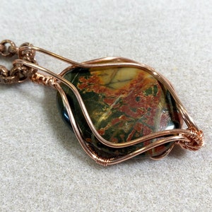 May include: A copper wire wrapped pendant featuring a green, brown, and red jasper stone. The pendant is attached to a copper chain.