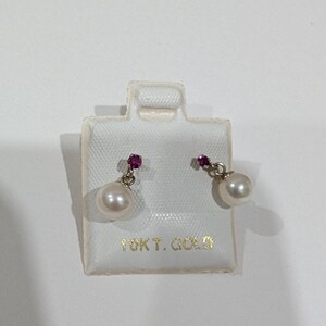May include: A pair of pearl earrings with a small red gemstone above each pearl. The earrings are on a white display card with the text "10K T. GOLD" printed in gold. The earrings are made of gold.