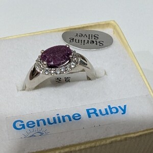 May include: A sterling silver ring with a genuine ruby gemstone. The ring features a large oval-shaped, dark pink ruby surrounded by small, clear accent stones. The ring is displayed in a box labeled "Genuine Ruby."