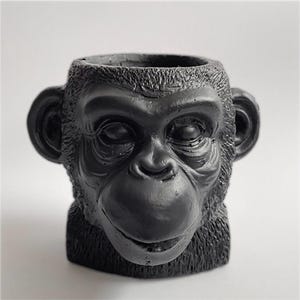Puede incluir: Una maceta negra con forma de mono con una superficie detallada y texturizada. La maceta tiene una amplia abertura para plantas y está diseñada para parecerse a la cara de un chimpancé, incluidas las orejas y los rasgos faciales.