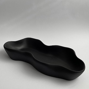 Könnte beinhalten: Eine mattschwarze Keramikschale mit einer länglichen, organischen und wellenförmigen Silhouette. Das Gefäß hat einen glatten, durchgehenden Rand, der sich nach innen und außen wölbt und eine fließende, asymmetrische Form mit einer tiefen Mulde bildet.
