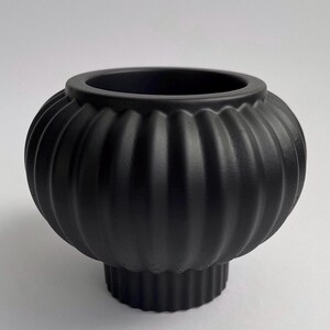 Può includere: Un vaso decorativo nero opaco con un design a coste, simile a una zucca. Il vaso ha un corpo arrotondato e una base cilindrica. L'apertura è di un colore scuro a contrasto. Un elemento di arredo casa moderno.
