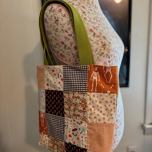 May include: A patchwork tote bag with a green strap. The bag features a variety of Halloween-themed fabric squares, including orange, black, and white patterns with skeletons, candy corn, and bats. The bag is displayed on a floral mannequin.