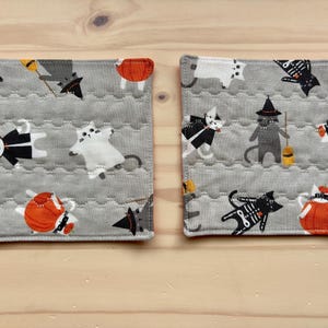 May include: Two grey, quilted fabric coasters with a Halloween cat theme. The coasters feature various cat designs dressed in costumes, including skeletons, witches, and pumpkins. The cats are in black, white, and grey, with orange accents.