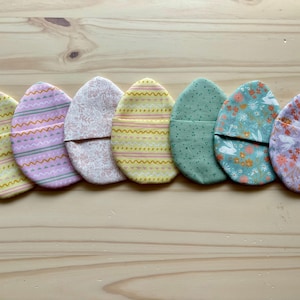 May include: Seven fabric Easter egg-shaped pouches in various pastel colors and patterns, including yellow, pink, green, and white. The pouches are lined up in a row on a wooden surface.