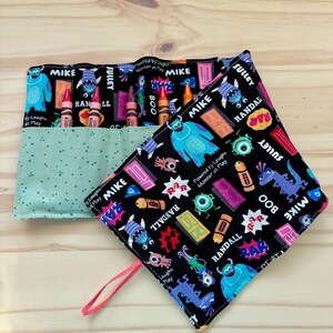 May include: A black and teal crayon roll and matching square pouch featuring colorful cartoon monster graphics and text. The crayon roll is open, revealing several crayons. The pouch has a pink loop for carrying.