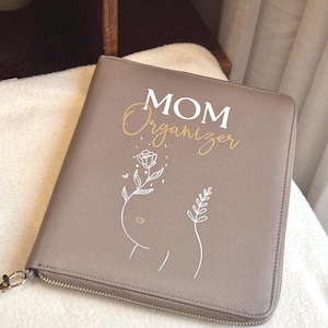 Könnte beinhalten: Ein taupefarbener Organizer mit Reißverschluss und den Worten "MOM Organizer" in Weiß und Gold. Das Cover zeigt ein minimalistisches Blumendesign mit einem Herzen. Der Organizer ist mit einem goldenen Reißverschluss und einem goldfarbenen Zipper verschlossen.