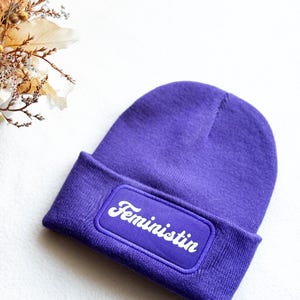Könnte beinhalten: Lilafarbene Mütze mit einem weißen Patch, auf dem "Feministin" in einer Retro-Schrift steht.
