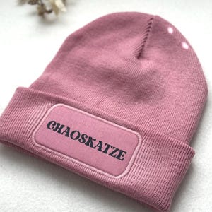 Könnte beinhalten: Eine altrosa Beanie-Mütze mit umgeschlagenem Saum. Die Mütze hat einen rechteckigen Aufnäher mit dem Wort "CHAOSKATZE" in schwarzer Schrift. Die Mütze ist aus weichem Strickmaterial.