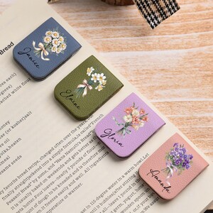Puede incluir: Cuatro marcapáginas rectangulares de cuero en azul, verde, morado y rosa, cada uno con un diseño floral y un nombre: Janice, Elaine, Gloria y Amanda. Están sobre una página de recetas.