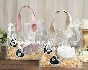 May include: Two clear plastic tote bags with daisy print and white ribbon bows. The bags are filled with shredded paper and have a heart-shaped object inside. The bags are personalized with the names "Sienna" and "Evelyn".