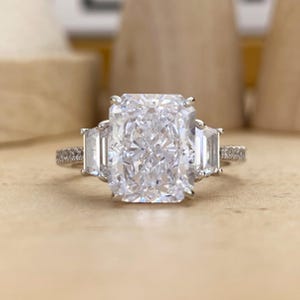 May include: A silver-toned ring featuring a large, rectangular radiant-cut center stone flanked by two smaller trapezoid-cut side stones. The thin band is adorned with a row of small, pavé-set stones along the sides, creating a brilliant, reflective appearance.