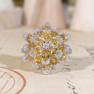 May include: A silver ring featuring a floral design. The ring is adorned with numerous yellow gemstones and small white gemstones, creating a sparkling, eye-catching effect. The ring's design resembles a flower.