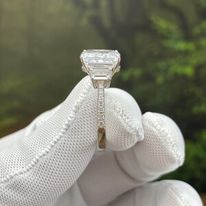 May include: A silver-toned ring featuring a large, rectangular, clear faceted center stone flanked by smaller rectangular side stones. The band is thin and encrusted with tiny, clear, round stones along the visible side, showcasing a polished metallic finish.