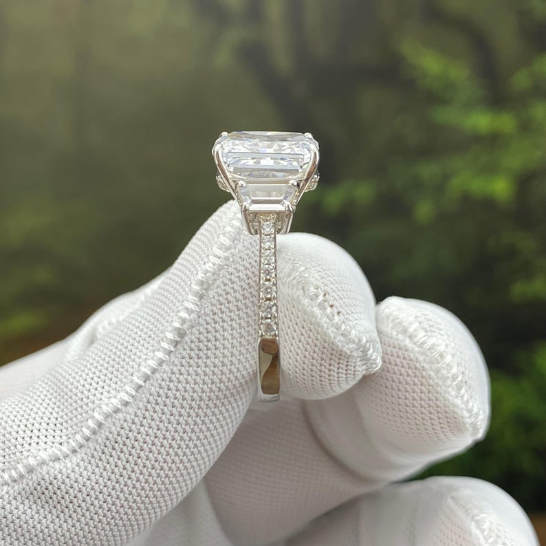 May include: A silver-toned ring featuring a large, rectangular, clear faceted center stone flanked by smaller rectangular side stones. The band is thin and encrusted with tiny, clear, round stones along the visible side, showcasing a polished metallic finish.