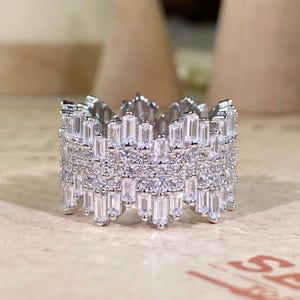May include: A silver-colored ring with a crown-like design. The ring is encrusted with numerous small, clear, rectangular and round-cut stones, creating a sparkling effect. The ring's design is intricate and detailed, with multiple layers of stones.