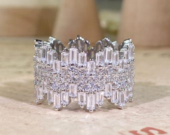May include: A silver-colored ring with a crown-like design. The ring is encrusted with numerous small, clear, rectangular and round-cut stones, creating a sparkling effect. The ring's design is intricate and detailed, with multiple layers of stones.