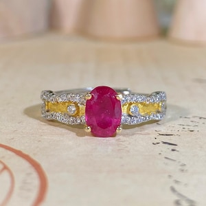 Puede incluir: Un anillo de dos tonos con una gran gema de rubí ovalada. El anillo tiene una banda plateada con detalles dorados y pequeñas gemas transparentes. El rubí es de color rojo vibrante y el anillo está sobre un fondo neutro.