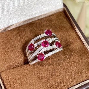 May include: A silver ring with multiple bands, each encrusted with small, clear stones. The ring features six vibrant, round, red gemstones. The ring is displayed in a brown jewelry box.
