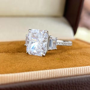 May include: A silver-toned ring featuring a large rectangular radiant-cut center stone flanked by two smaller trapezoid-cut side stones. The band is crafted from a metallic material and is accented with small pav&eacute;-set stones along the visible shank.