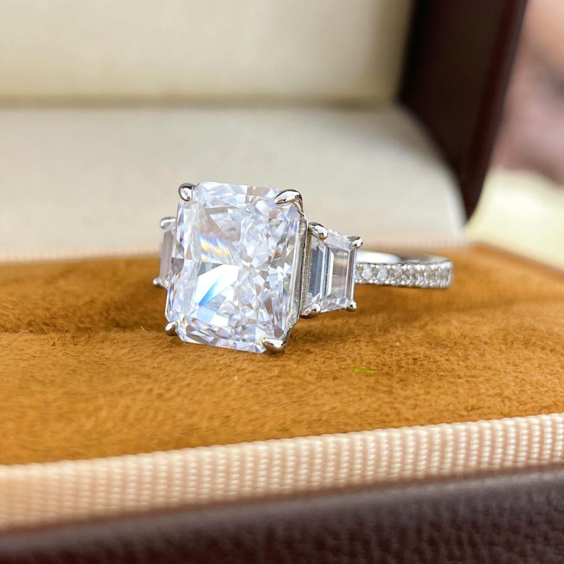 May include: A silver-toned ring featuring a large rectangular radiant-cut center stone flanked by two smaller trapezoid-cut side stones. The band is crafted from a metallic material and is accented with small pav&eacute;-set stones along the visible shank.
