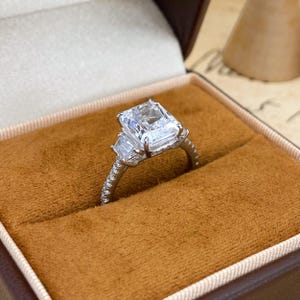 May include: A silver-toned ring featuring a large emerald-cut center stone flanked by two smaller baguette-cut stones on each side. The thin band is encrusted with small, round, clear stones along the shank, set in a metallic frame.