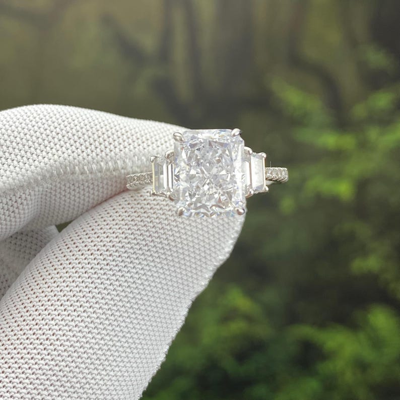 May include: A silver-toned ring featuring a large rectangular radiant-cut center stone flanked by two smaller baguette-cut side stones. The band is thin and encrusted with small pav&eacute;-set stones along the visible portion of the metal shank.