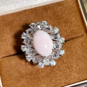May include: A ring featuring a large, smooth, oval-shaped pale pink cabochon stone at the centre, surrounded by a halo of multiple clear, faceted, pear-shaped and round white stones set in a silver-toned metal frame with a polished finish.