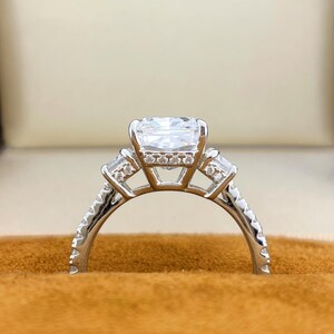 May include: A silver-toned ring featuring a large rectangular center stone, flanked by two smaller baguette-cut stones on each side. The band and the gallery beneath the center stone are adorned with small, clear, round-cut accent stones.