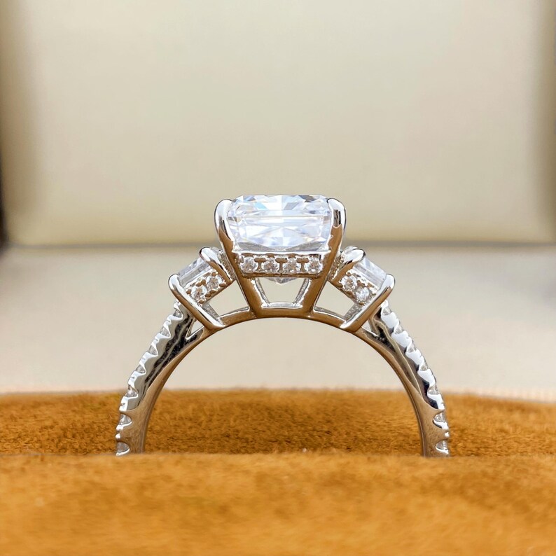 May include: A silver-toned ring featuring a large rectangular center stone, flanked by two smaller baguette-cut stones on each side. The band and the gallery beneath the center stone are adorned with small, clear, round-cut accent stones.